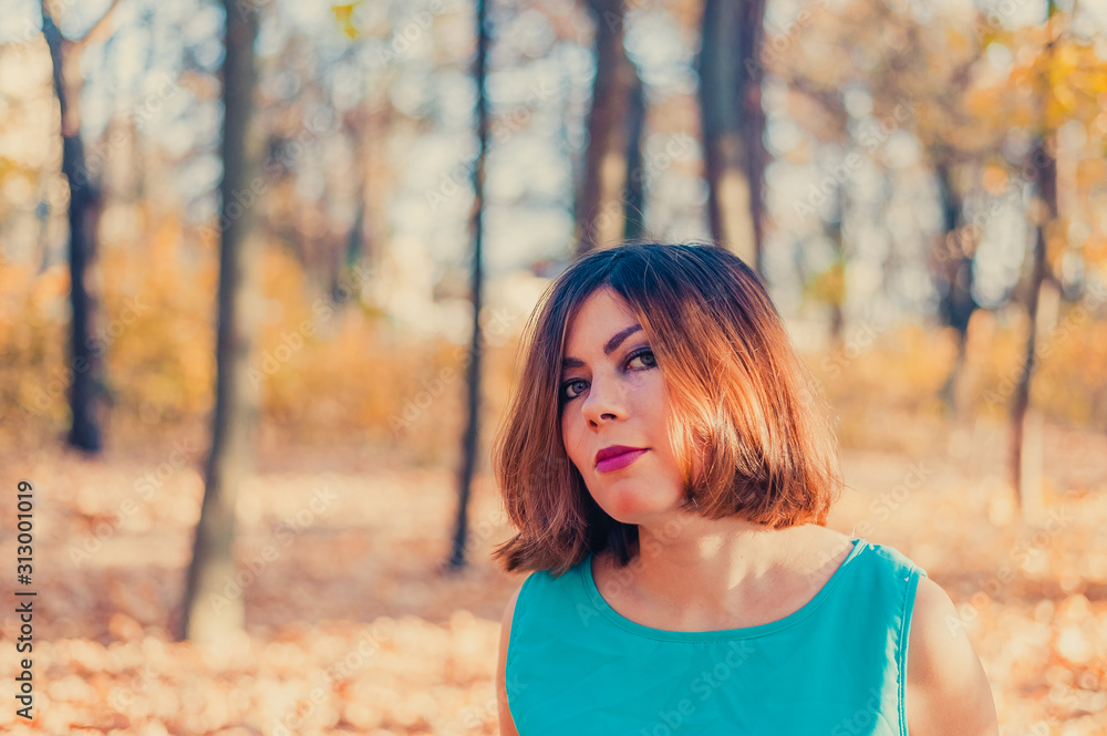 cute young red-haired girl with beautiful eyes in a blue dress looks at the camera in the autumn yellow forest