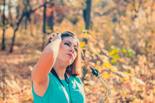 very cool young brunette girl with makeup in a blue dress with a saxophone in her hands straightens her hair, standing in a yellow autumn forest