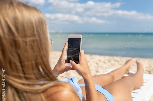 Back view a beautiful girl in a blue swimsuit is using a smartphone and sunbathing on a sun lounger near the beach with a blue sea