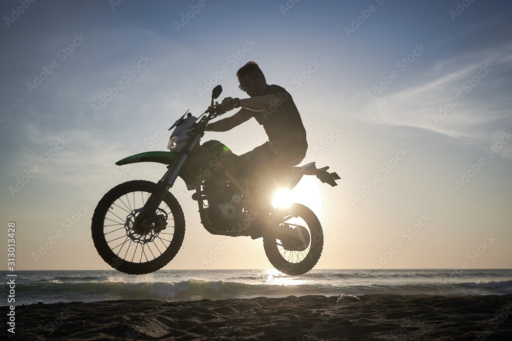 Fototapeta premium Man on the motorbike mke jump on the black sand beach against sun
