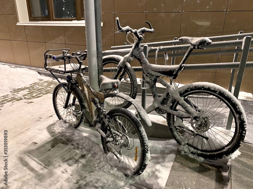 Two snow-covered bikes parked with a tether near a pillar near the ramp near the entrance of the house entrance.