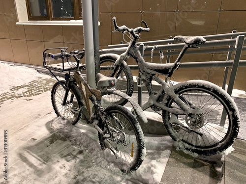 Two snow-covered bikes parked with a tether near a pillar near the ramp near the entrance of the house entrance.