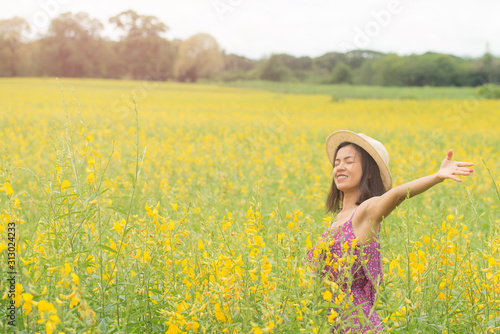 asia woman with a hat in her hand walks in a field with field flowers and smiles sincerely, happy enjoying summer in yellow field at sunset. smiling with arms raised up. concept of freedom. © nareekarn