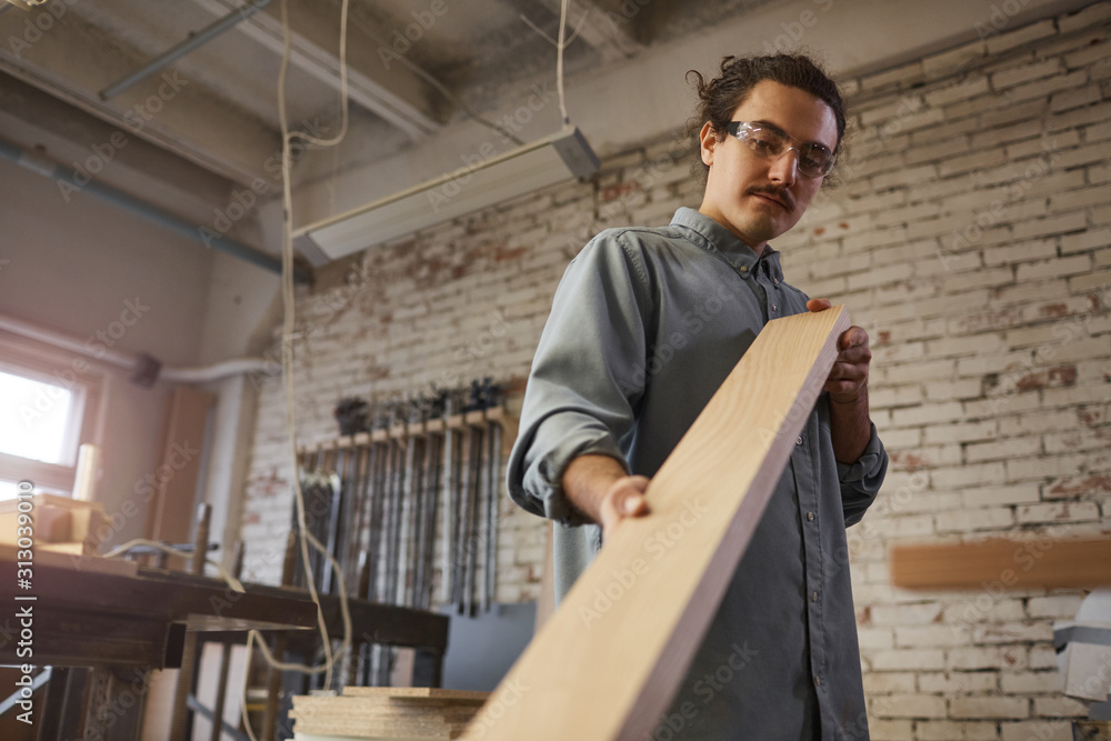 Young serious worker examining wooden detail while working at carpentry industry