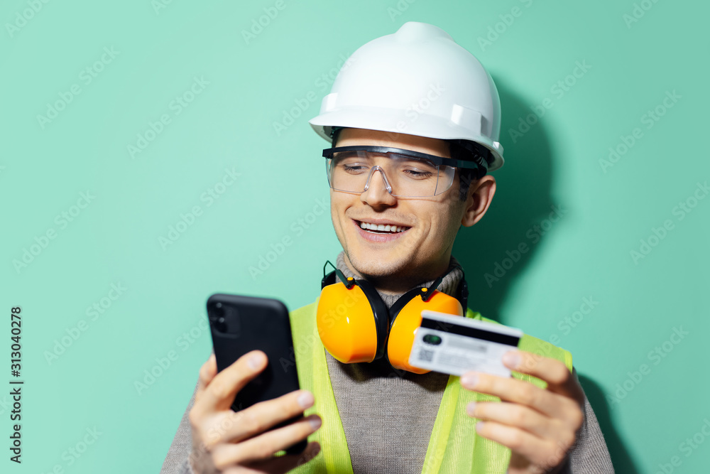 Studio portrait of happy young builder man, engineer, paying online with smartphone and credit card. Wearing construction safety helmet, glasses and headphones on background of aqua menthe color. Studio portrait of happy young builder man, engineer, paying online with smartphone and credit card. Wearing construction safety helmet, glasses and headphones on background of aqua menthe color.
