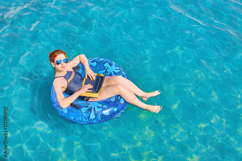 Cute young redhead woman with sunglasses in inflatable ring relaxing in the sea with laptop. Freelancer concept, remote job