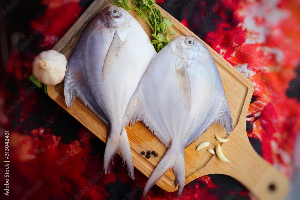 White Pomfret fish or butter fish places on a wooden board with garlic cloves, black pepper seeds and coriander