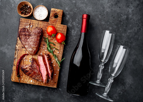two grilled beef steaks in the form of a heart with spices, a bottle of champagne or wine with two glasses for dinner for Valentine's day on a stone background with copy space for your text