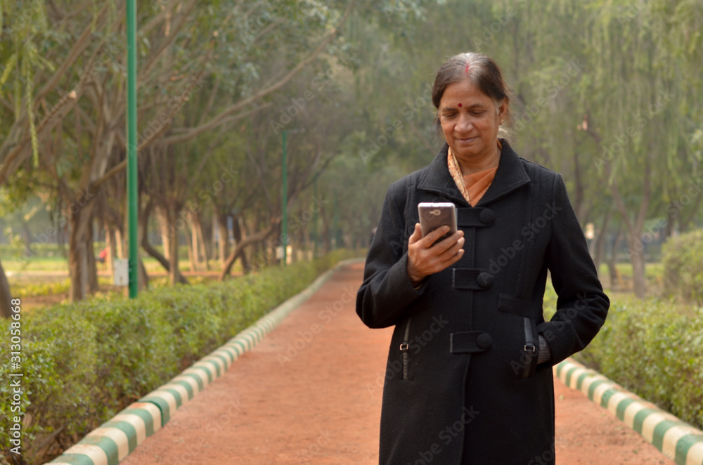 Indian woman working on a mobile phone, experimenting with technology in a park during winters in Delhi. Woman wears a saree and a black coat. Concept shot showcasing Digital Literacy in India Indian woman working on a mobile phone, experimenting with technology in a park during winters in Delhi. Woman wears a saree and a black coat. Concept shot showcasing Digital Literacy in India