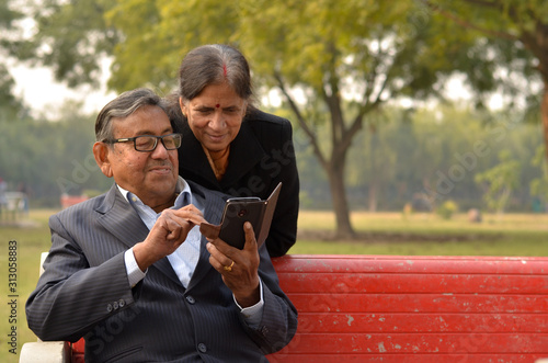 Wallpaper Mural Portrait of a happy looking retired senior smart Indian man and woman couple smiling, talking and looking at mobile phone screen while sitting on a park bench in New Delhi, India. Concept love Torontodigital.ca