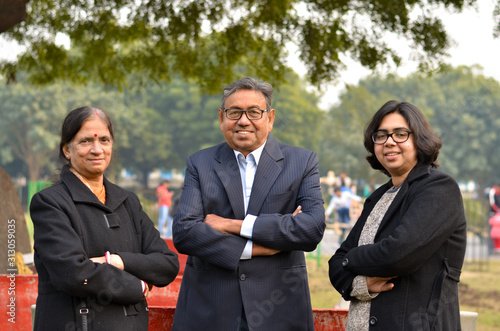 Portrait of a happy Young Indian woman with her mother and father wearing blazers and over coats in winters standing with crossed hands in a park in looking at the camera. Successful business family