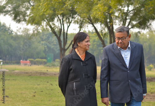 Carefree & happy elder man in black suit, walking with his wife who is wearing black coat walking in a park in winter in New Delhi, India. Concept love & togetherness