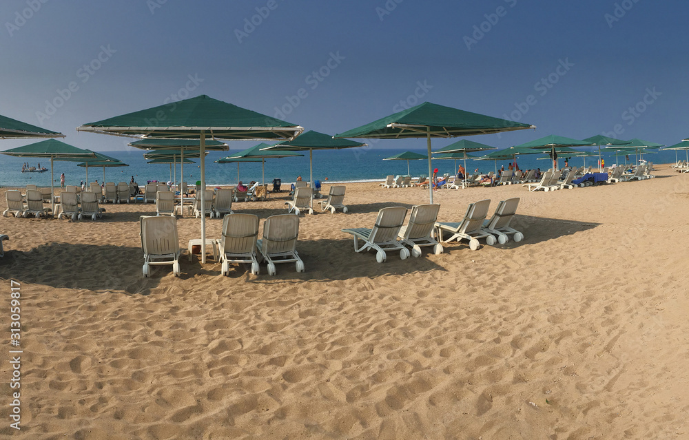 Panoramic view of a yellow sandy beach with white sunbeds in several rows, green fabric awnings, blue sea and dark blue sky with no clouds. Travel concept. Panoramic view of a yellow sandy beach with white sunbeds in several rows, green fabric awnings, blue sea and dark blue sky with no clouds. Travel concept.