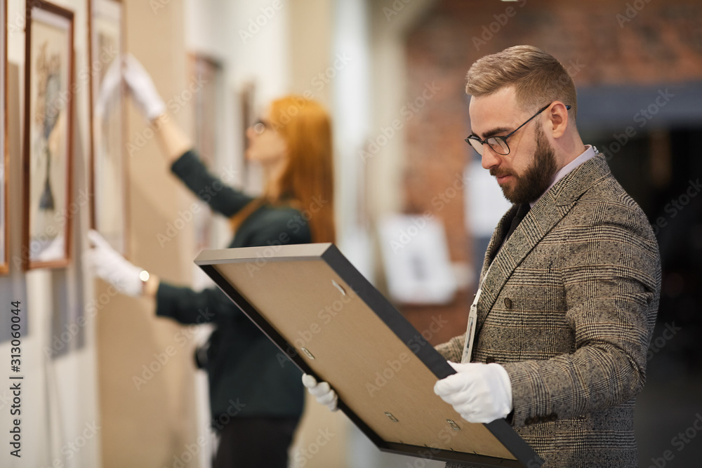 Art manager in suit and in gloves standing an looking at picture in his hands while woman hanging painting on the wall at gallery Art manager in suit and in gloves standing an looking at picture in his hands while woman hanging painting on the wall at gallery
