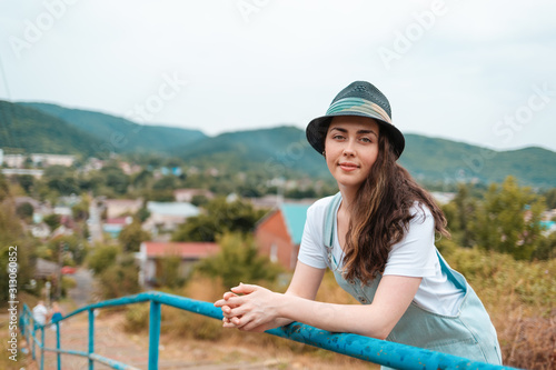 Portrait of a young beautiful brunette woman in hat posing near the railing against a rural view
