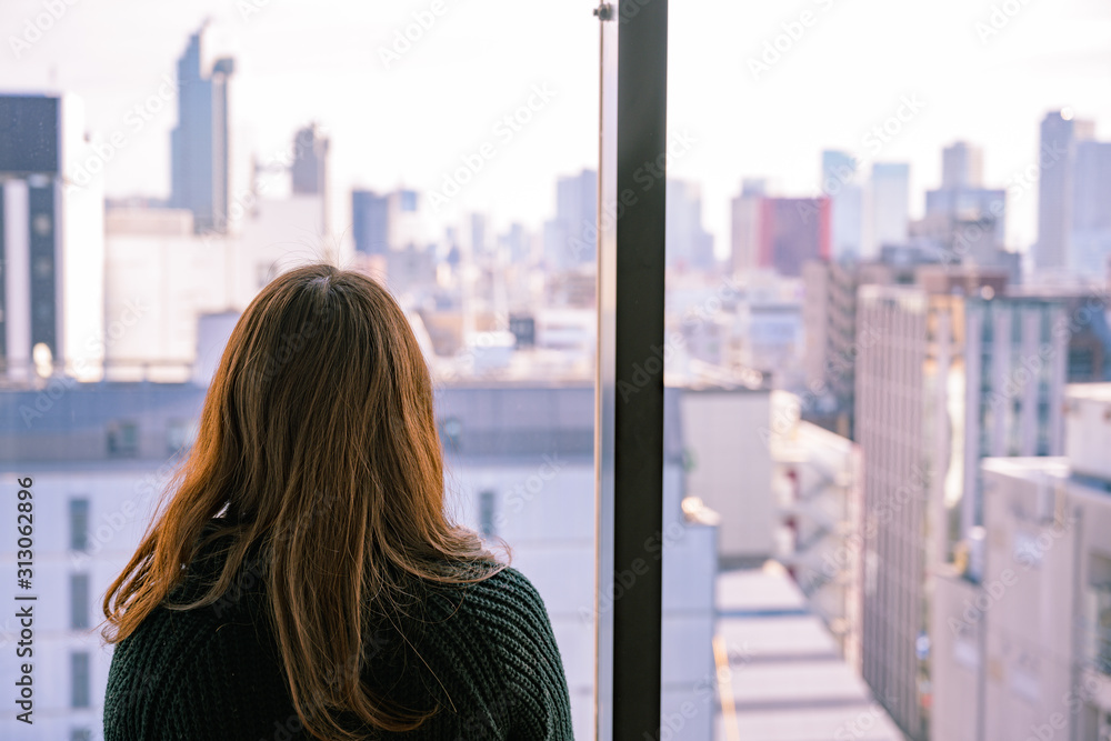 高層ビルの窓から都会の景色を眺める若い女性の後ろ姿