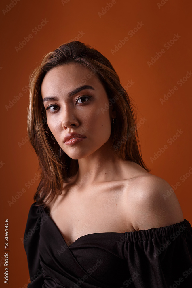 charming elegant fashion model wearing black dress with deep neckline posing on orange background. asian skinny young woman sits in sexy evening gown. beautiful sensual glamorous girl poses in studio charming elegant fashion model wearing black dress with deep neckline posing on orange background. asian skinny young woman sits in sexy evening gown. beautiful sensual glamorous girl poses in studio