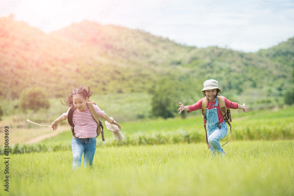Fototapeta premium Two children carrying a backpack in their backs and wearing a hat, standing under a tree in the countryside. She walked and enjoyed the surrounding nature. She enjoys traveling during the summer.