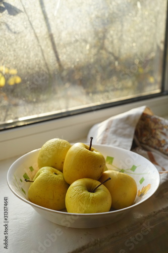 Natural yellow apples in a white plate on a windowsill, organic fruits on a sunny winter day.