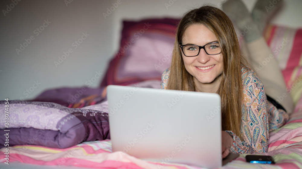 Junge Frau mit Laptop liegt zuhause auf dem Bett und lächelt in die Kamera Junge Frau mit Laptop liegt zuhause auf dem Bett und lächelt in die Kamera