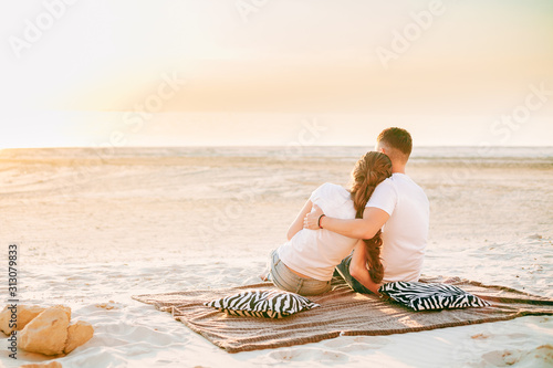 A guy and a girl on the beach at beautiful sunset sit on a plaid and gently hug each other and look into the distance. Holiday concept - Valentine's Day, love on the beach, lovers, romance, couple.