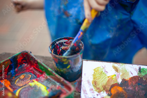 Child at nursery painting with a brush wearing a painting apron with paint all over it. Close up of the paints being mixed in a cup to make new colours for the picture they are making. 