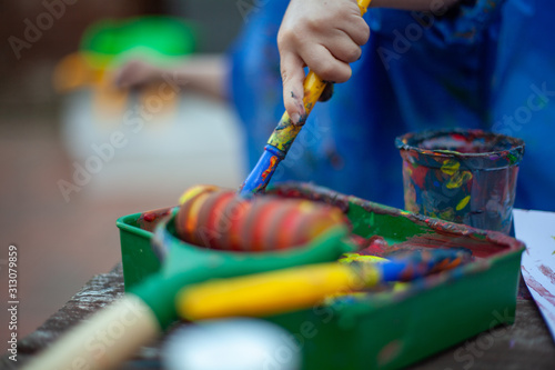 Child at nursery painting with a brush and roller, learning colour and creativity through messy play. Close up of the paints and the child holding a paint brush to create a painting.