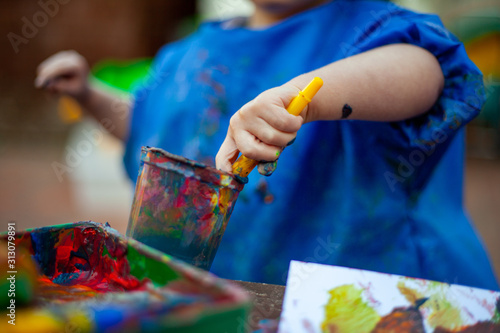 Child at nursery painting with a brush wearing a painting apron with paint all over it. Close up of the paints being mixed in a cup to make new colours for the picture they are making. 