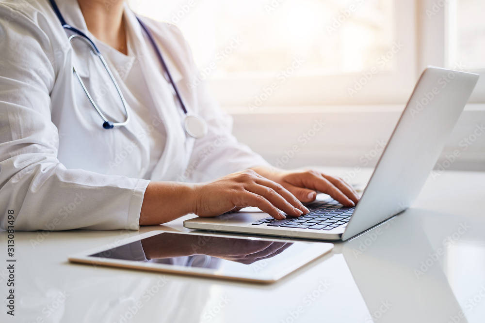 Female doctor typing on her laptop computer in medical office - obrazy, fototapety, plakaty 
