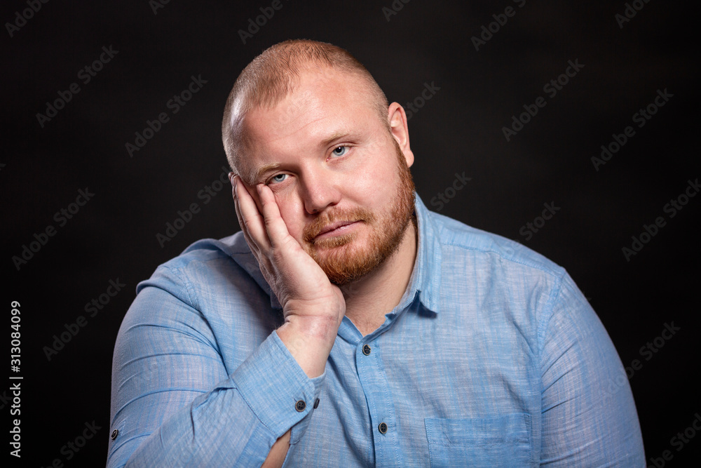 Fat red-haired man with a beard and mustache in a blue shirt is sad, propping his head in his hands. Black background. Close-up.