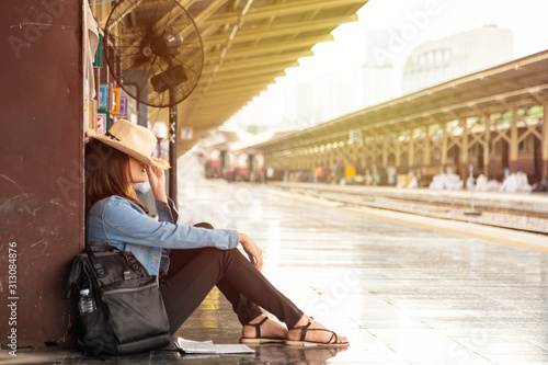 Beautiful Asian woman in a white T-shirt hiding face under straw hat from sunlight, She is sitting on floor learning a brown pillar with a black fan at train station