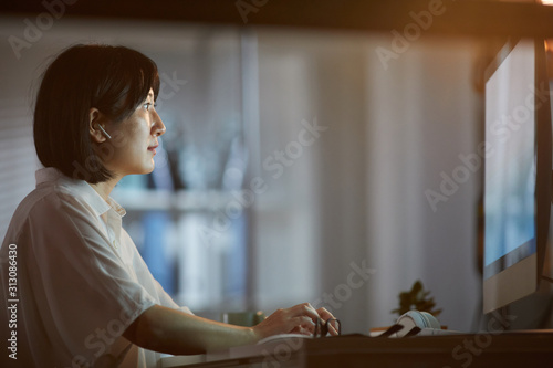 Side view of Asian woman with dark short hair sitting in front of computer monitor and typing in dark office