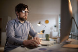 Serious bearded businessman in eyeglasses looking at computer monitor while sitting at his workplace at office