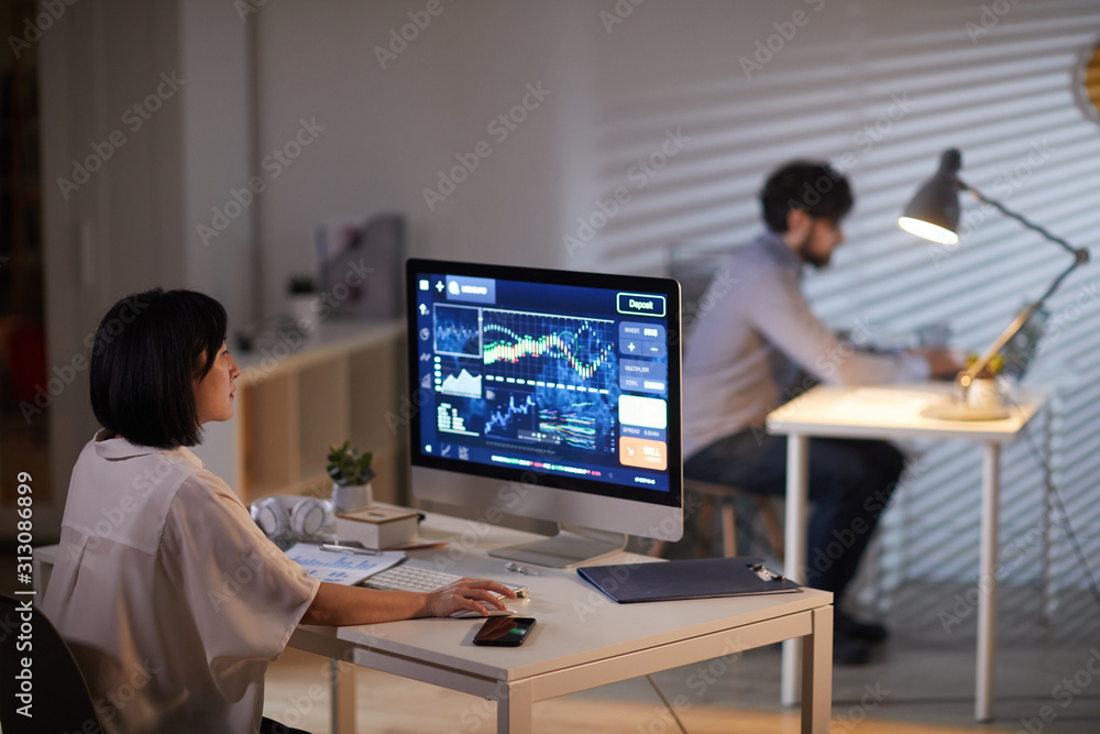 Rear view of young businesswoman sitting at her workplace and working online with financial report with businessman working at the table in the background
