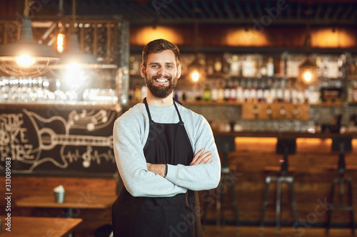 Bearded smiling barman waiter standing on the background of a bar.