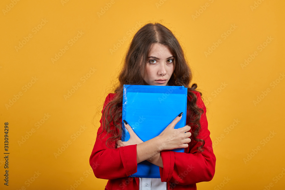 Serious brunette young woman in fashion white shirt and red jacket keeping blue folder, looking at camera isolated on orange background in studio. People sincere emotions, lifestyle concept. Serious brunette young woman in fashion white shirt and red jacket keeping blue folder, looking at camera isolated on orange background in studio. People sincere emotions, lifestyle concept.