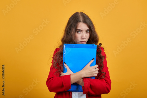 Serious brunette young woman in fashion white shirt and red jacket keeping blue folder, looking at camera isolated on orange background in studio. People sincere emotions, lifestyle concept.
