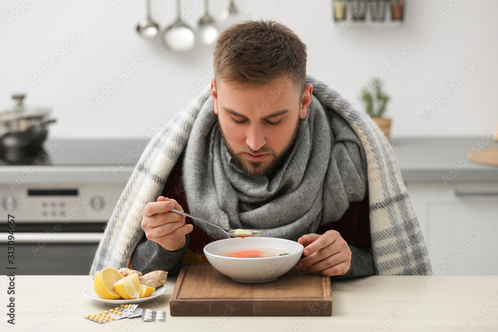 Sick young man eating tasty soup to cure flu at table in kitchen Sick young man eating tasty soup to cure flu at table in kitchen