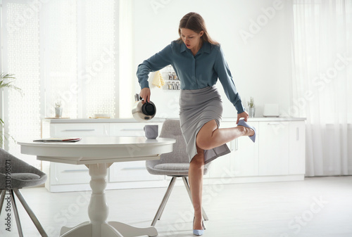 Young woman pouring coffee into cup while putting on shoes at home in morning photo