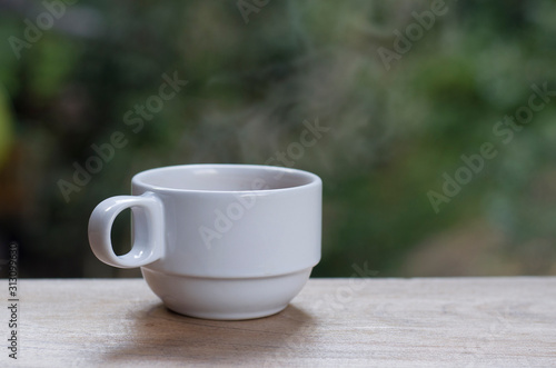 White hot coffee cup with smoke floating out Put on a wooden balcony With a blurred tree backdrop