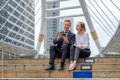 two businessmen sit looking on mobile phone in front of office building