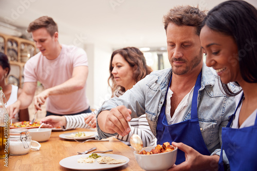 Group Of Men And Women Sitting Around Table Eating Meal They Have Prepared In Kitchen Cookery Class