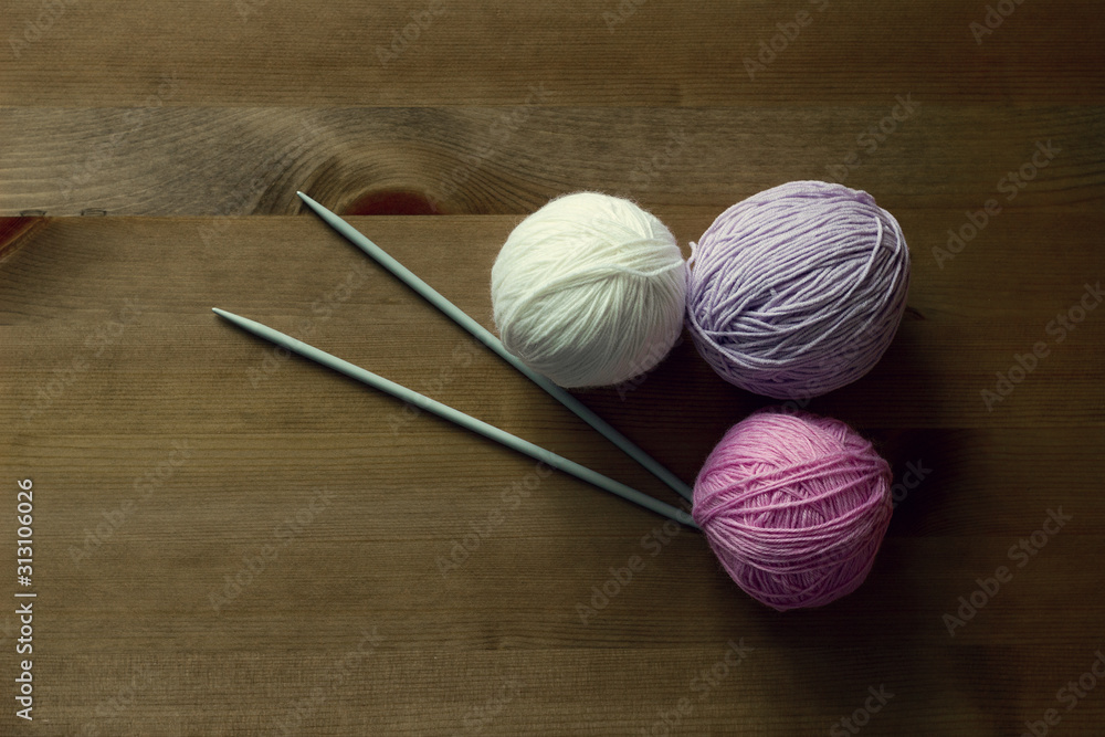 Balls of white, lilac and pink yarn and two metal knitting needles on a wooden table
