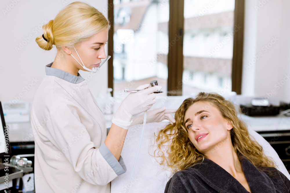 Obraz premium Young woman cosmetologyst making professional facial hydration to a pretty woman at the beauty spa clinic, close-up view
