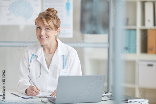 Portrait of mature female doctor sitting at the table in front of laptop and smiling at camera while filling the documents at office