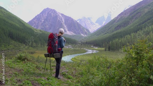 A young girl traveler with a backpack enjoys a mountain river and snowy peaks on a summer hike. Lifestyle concept relax in outdoor. 4k footage.