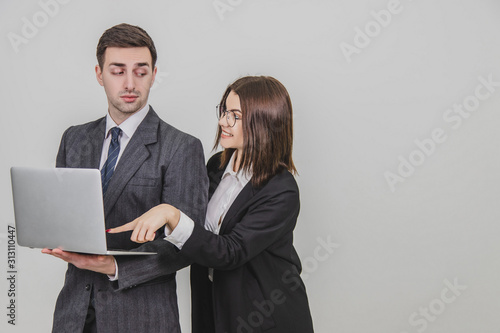 Handsome man standing, working with laptop. Woman is pointing from man's behind at something in the laptop, interfering him. Man is annoyed because of this.