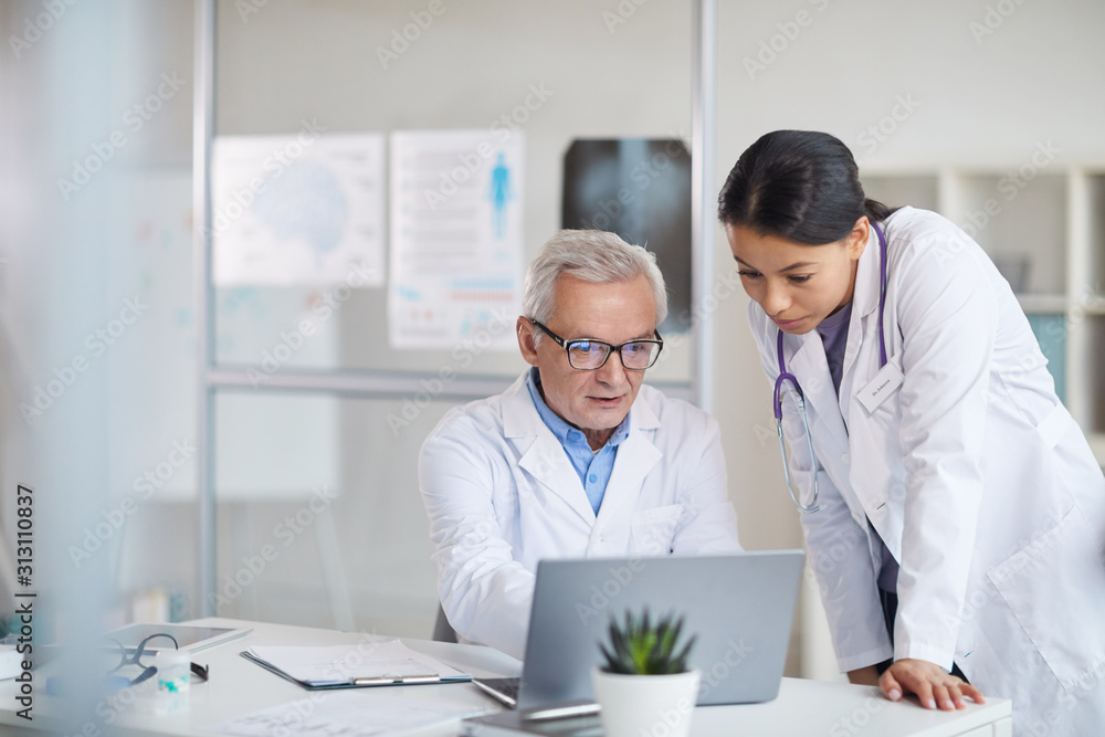 Senior doctor in white coat sitting at the table working on laptop and discussing work with his nurse at office
