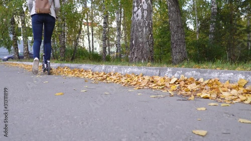 Close-up girl rides on a scooter on an asphalted track covered with autumn leaves. White sneakers and jeans - casual style. 4k footage.