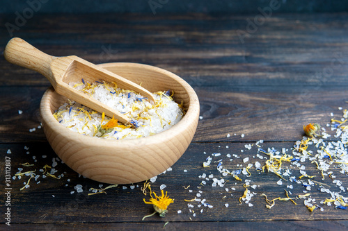 White fragrant sea salt crystals with dried calendula flowers in wooden bowl with spoon on dark rustic wooden background. Spa, body and health care concept. Copy space.
